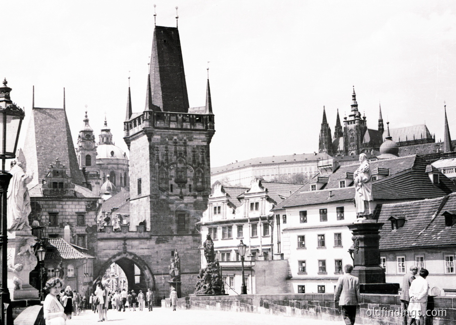 Black-and-white shot of Prague’s Charles Bridge (Karlův most) with Gothic-style statues and the Old Town Tower (14th-century) dominating the center. Crowds of pedestrians traverse the arched bridge, flanked by historic buildings and spires. Architectural details include ornate stonework and traditional Czech rooftops.