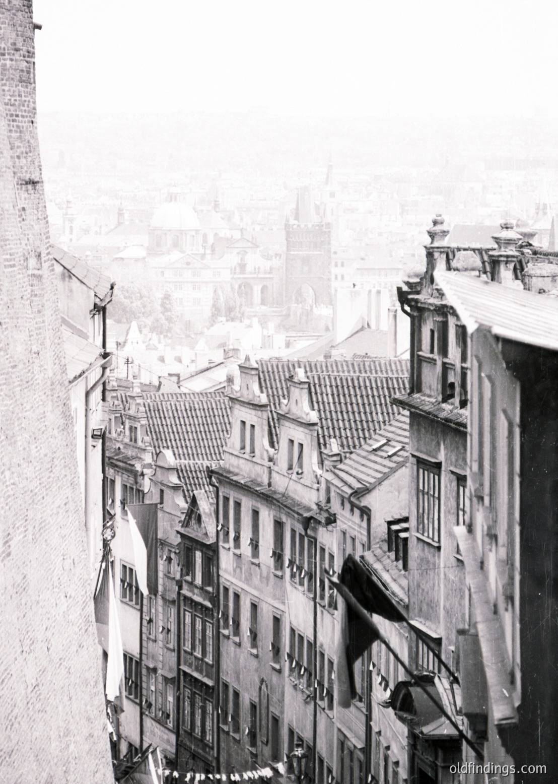Black-and-white aerial view of a European urban alleyway, likely 19th–early 20th century. Narrow, multi-story buildings with tiled roofs, wrought-iron balconies, and hanging laundry. Overhead wires crisscross above, and a flagpole with a flag (partially visible) suggests civic or national significance. Dense, historic architecture hints at a preserved old town.