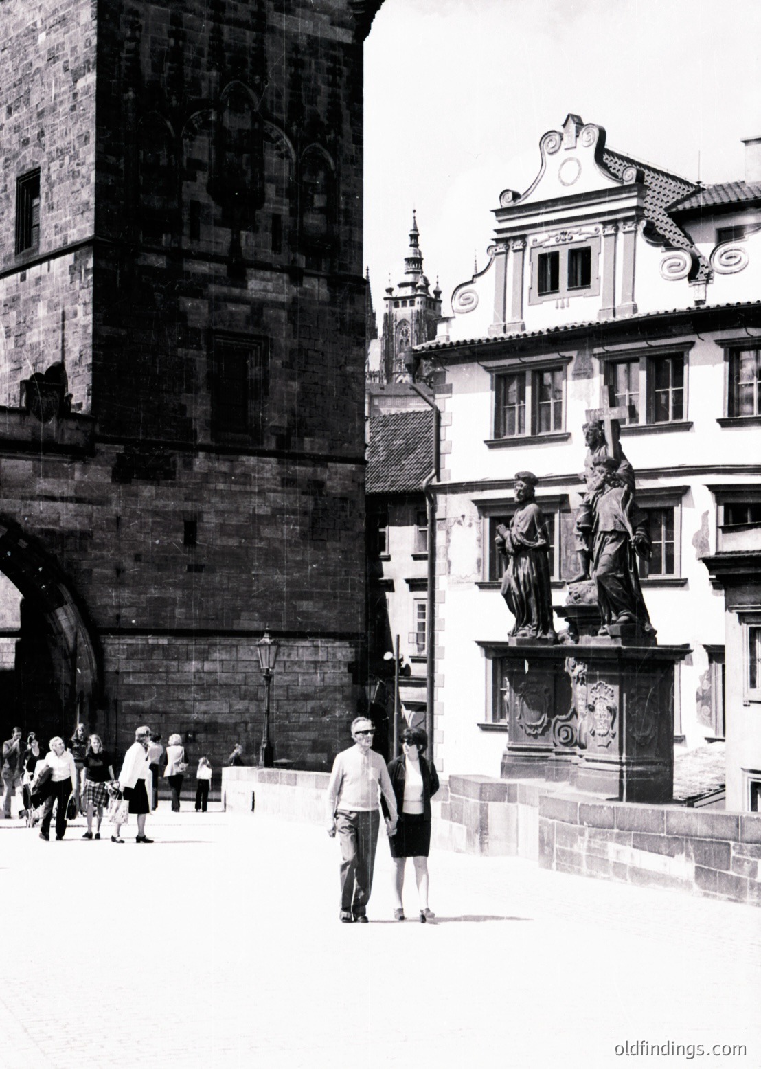 Historic Prague courtyard featuring Gothic stone archway and Baroque-style statue. Two figures in mid-20th-century attire (1950s–60s) pose near a pedestal adorned with classical reliefs. Crowd of tourists explores the Old Town’s architectural blend.