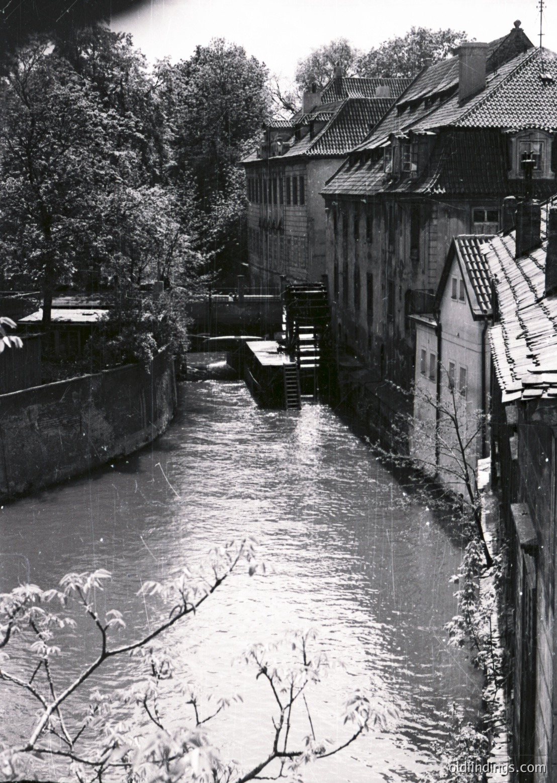 Historic waterwheel along a narrow canal flanked by timber-framed buildings with steeply pitched roofs, likely European . Stone bridge and lush greenery frame the scene, evoking industrial-era architecture. Ideal for vintage travel or design references.