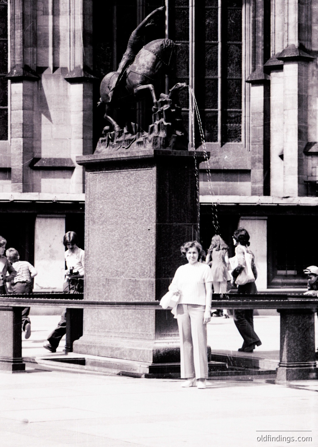 Neoclassical equestrian statue atop a stone pedestal in an urban plaza, surrounded by classical architecture. A woman in 1970s-style wide-leg trousers and blouse poses near the base. Crowd of casually dressed pedestrians in mid-20th-century attire.