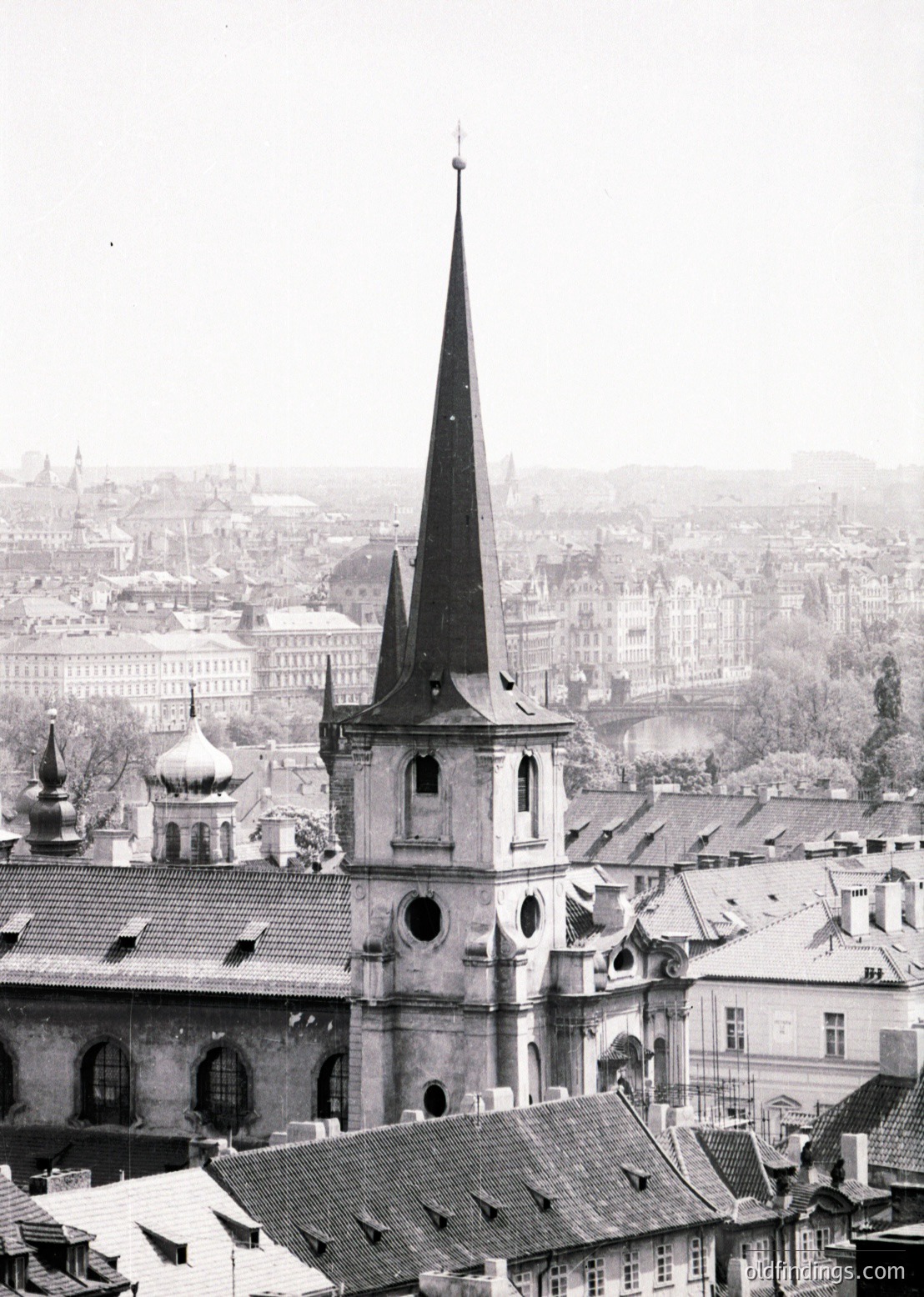 Prominent Gothic Revival church spire dominates Prague’s historic skyline, framed by Baroque-style rooftops below. Mid-19th century architecture contrasts with distant 20th-century buildings.