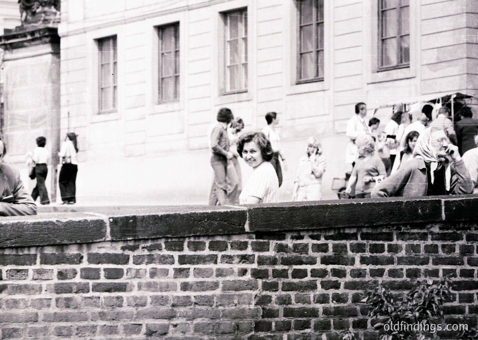Black-and-white snapshot of a lively courtyard scene, likely 1960s–1970s. A group of people—mostly young—gather around a brick wall, some standing, others seated. The background features a large, institutional-style building with tall windows. Casual, candid poses suggest a social or educational setting.