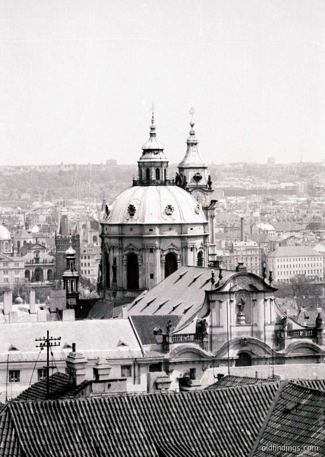 Baroque-style dome with twin spires dominates rooftops of a densely built European cityscape. Snow covers rooftops, indicating winter. Urban architecture blends historic and utilitarian styles. Likely , , .