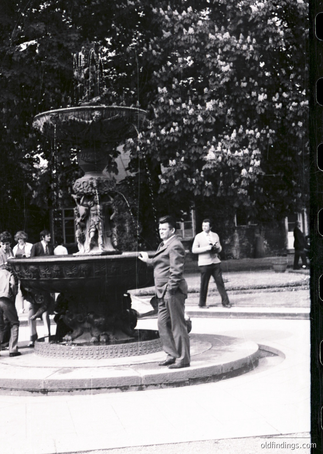 Classic 1960s fountain scene with ornate multi-tiered basin and sculpted figures. Central man in suit poses near water spout, surrounded by casually dressed onlookers in a tree-lined plaza.