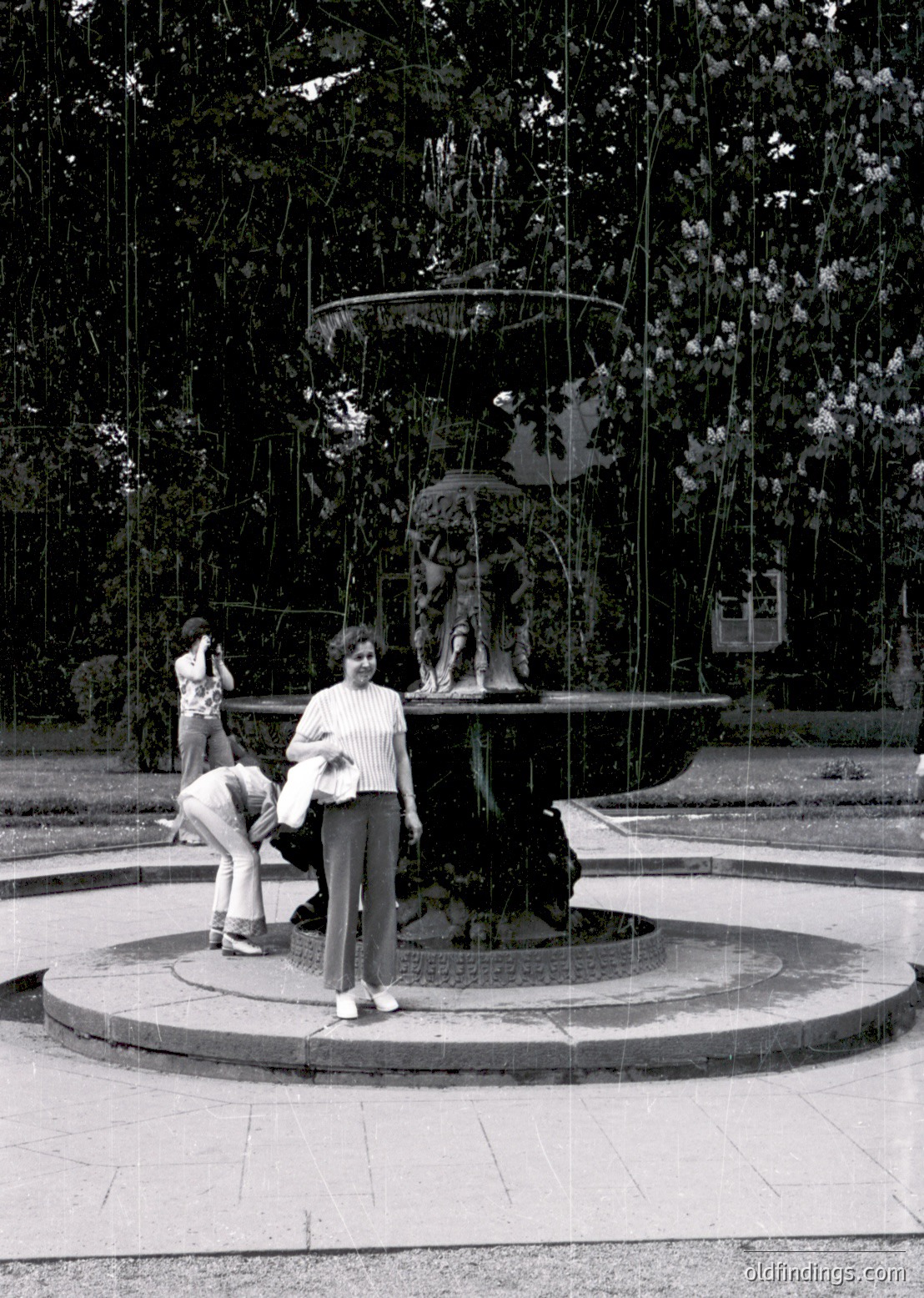 Classic 1970s-style fountain with sculpted figures and cascading water in a park setting. Two women pose near the center, one in a striped sweater and wide-legged trousers. Lush greenery and trees frame the scene, suggesting a European urban park. Black-and-white film captures mid-century urban life.
