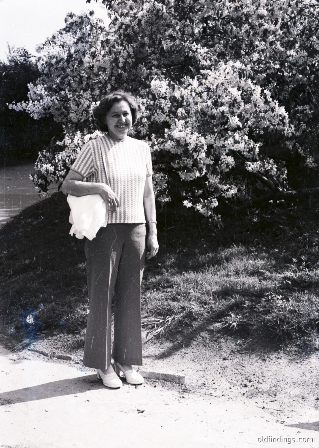 Mid-century woman in a striped sweater and wide-leg trousers stands outdoors, holding a rolled document or fabric. Lush greenery and a paved path frame the scene, suggesting a park or garden setting. Style hints at 1960s–1970s Western Europe or North America.