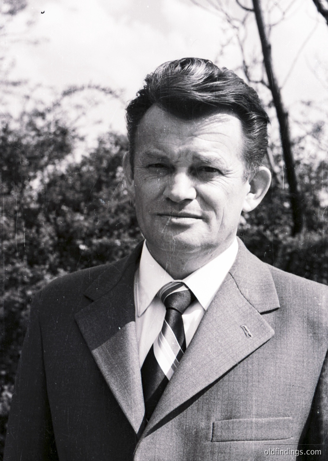 Portrait of a man in a sharp 1960s-70s suit, tie, and slicked-back hairstyle against an out-of-focus forest backdrop. Formal attire suggests professional or official context.