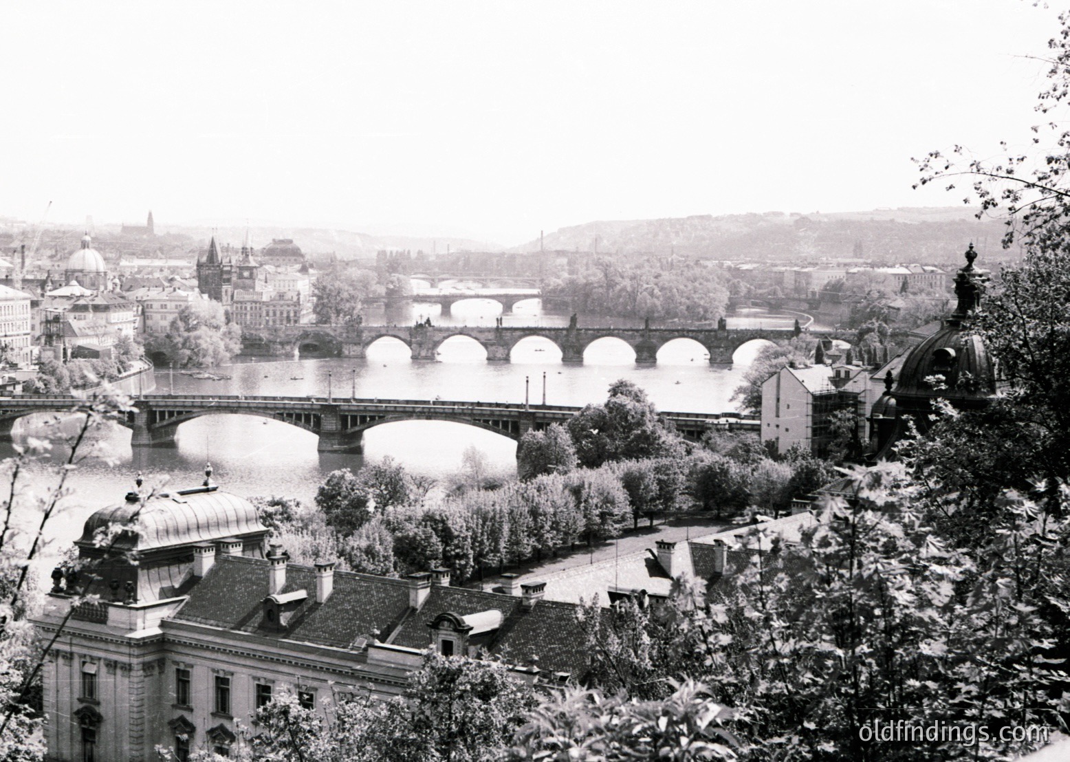 Black-and-white view of Prague’s Charles Bridge spanning the Vltava River, flanked by historic Baroque architecture. The bridge’s Gothic towers and arcades dominate the scene, with Prague Castle’s silhouette visible in the background. Likely late 19th to early 20th century.