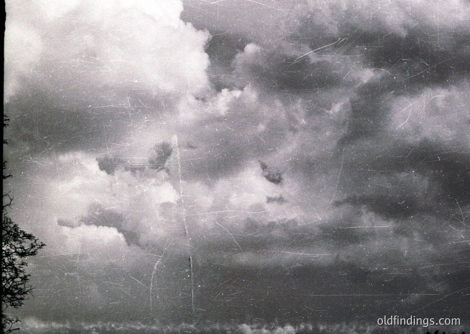Vintage sepia-toned photograph of stormy skies with dramatic, billowing clouds and visible scratches/tears. Partial tree silhouette at lower left suggests outdoor setting. Likely early-to-mid 20th century due to photographic style.