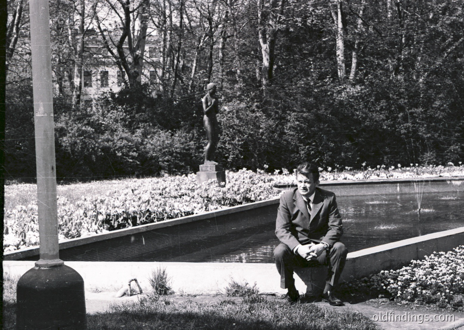 Mid-20th century man in formal attire sits by a reflective water feature in a landscaped park. Classical statue and manicured greenery in background suggest European urban park design. Likely 1950s–1960s.