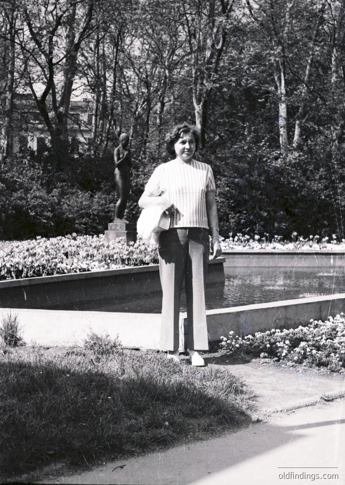Black-and-white portrait of a woman in a 1970s park setting, holding a white bag. She wears a striped blouse, wide-leg trousers, and white sneakers. A modernist bronze sculpture of a standing figure stands behind her. Lush greenery, flower beds, and a paved pathway frame the scene.