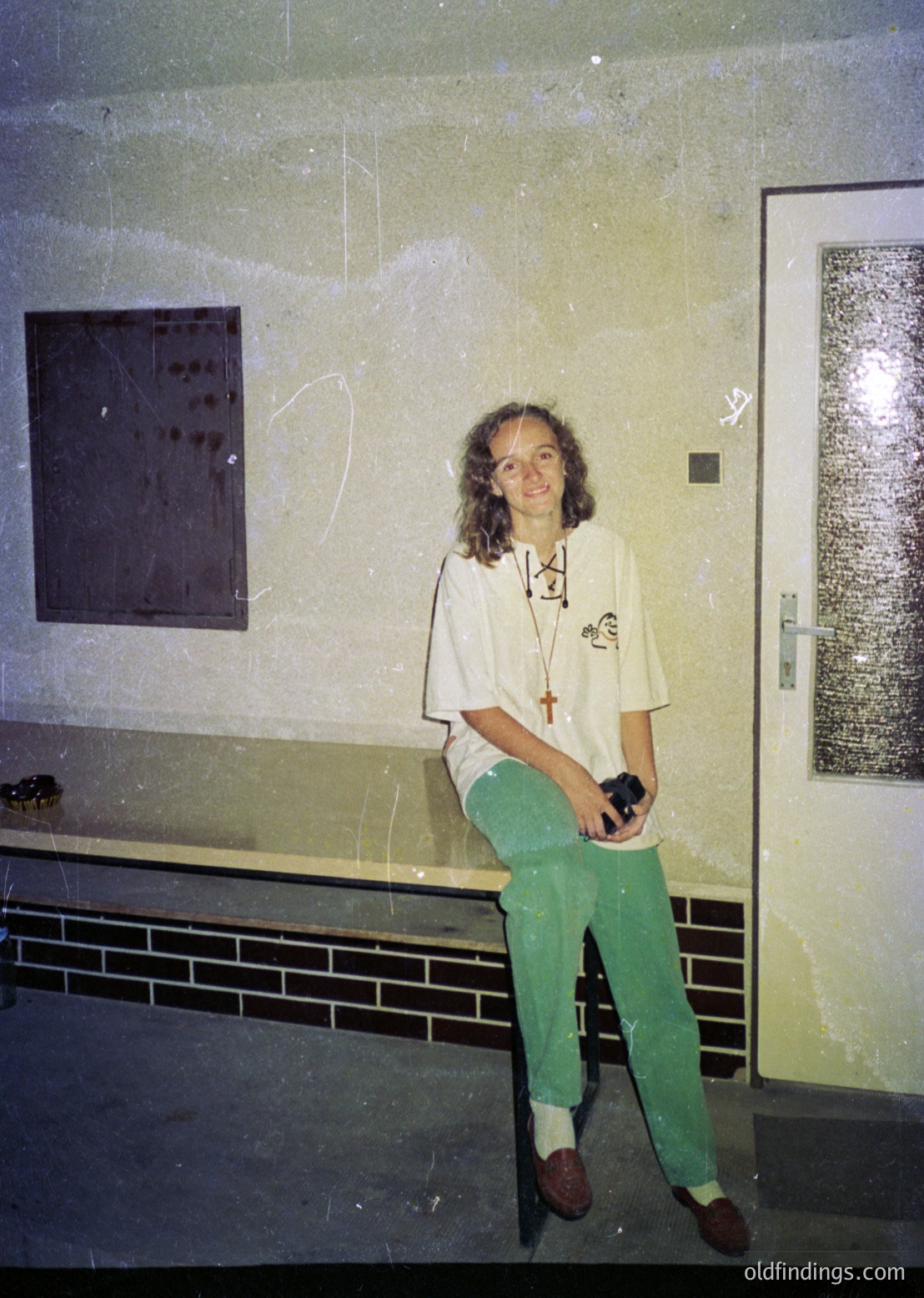 Indoor portrait of a person in a utilitarian space, likely a 1970s–1980s institutional or medical setting. The individual wears a white long-sleeve shirt with a lanyard (medical ID or badge), green pants, and brown shoes. A cross necklace and camera suggest a blend of professional and personal interests. Plain walls, tiled baseboards, and a door with a metal mesh panel indicate a functional, possibly clinical environment.