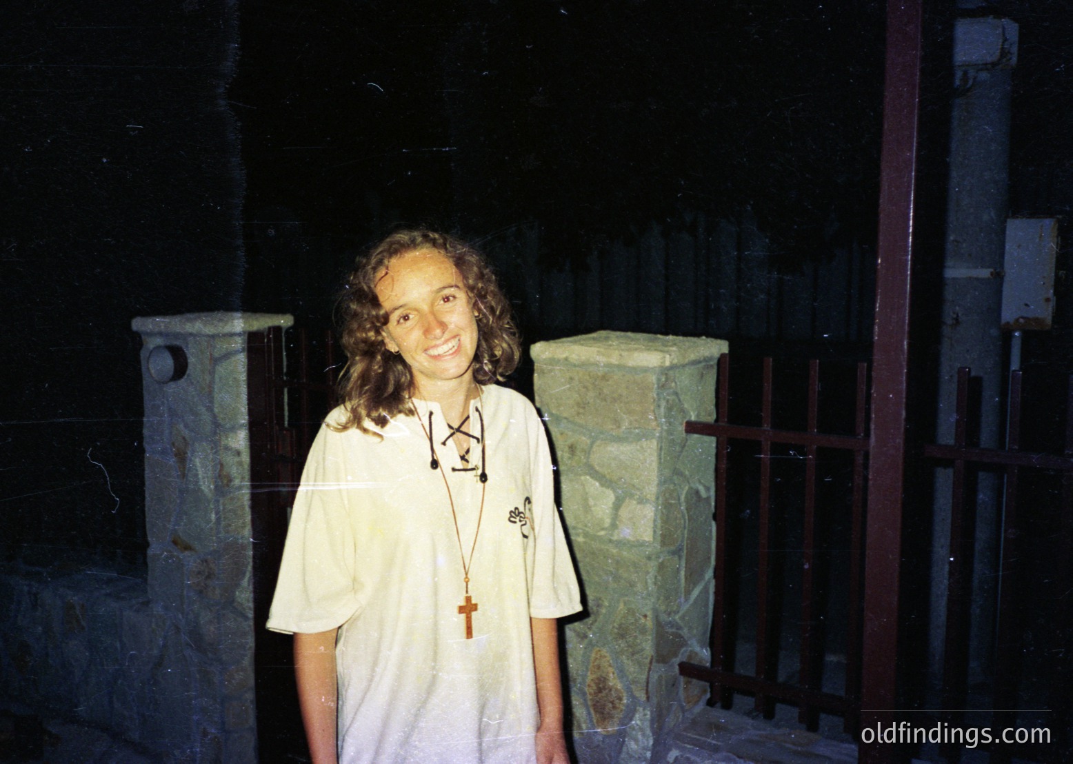 Young woman in a white shirt with a lanyard featuring a cross pendant, standing indoors near stone columns with intricate carvings. Likely a religious or historical site.