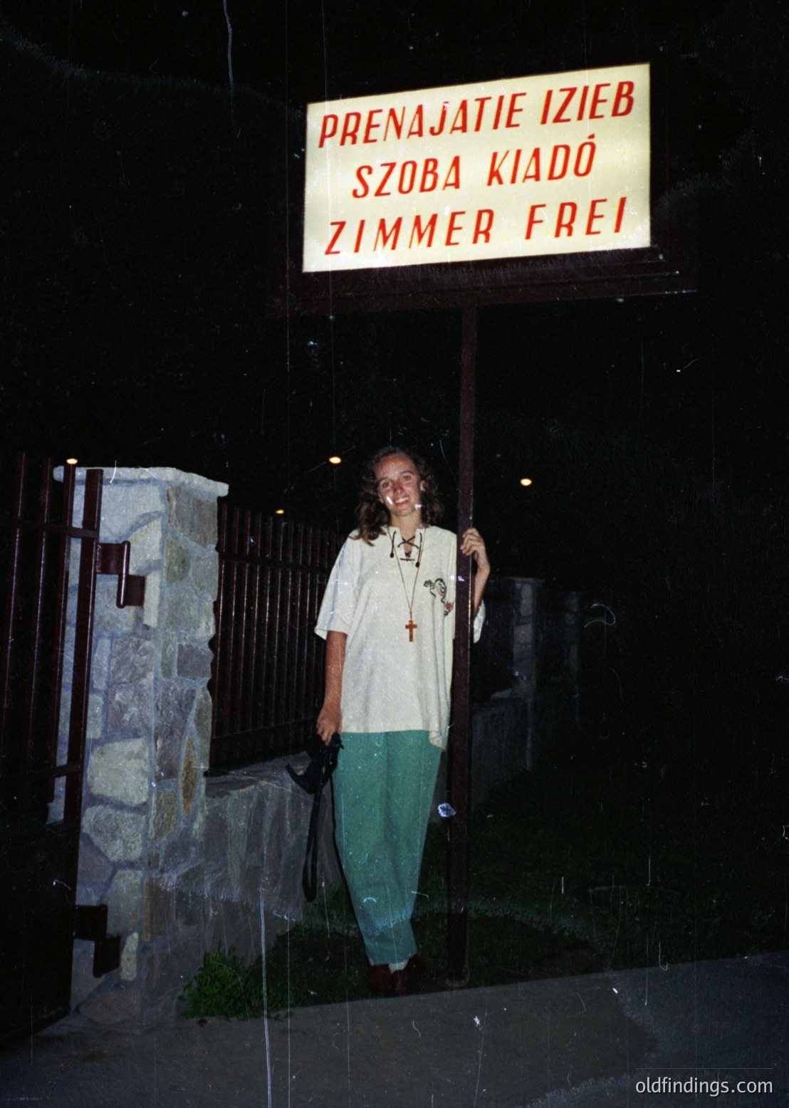 Vintage sign in Polish/Romanian/German ("Prenajatie Izieb Szoba Kiadó Zimmer Frei") outside a building, indicating available rooms for rent. Woman in 1970s-style clothing (white top, green pants) poses near entrance. Reflective glass door suggests urban setting.