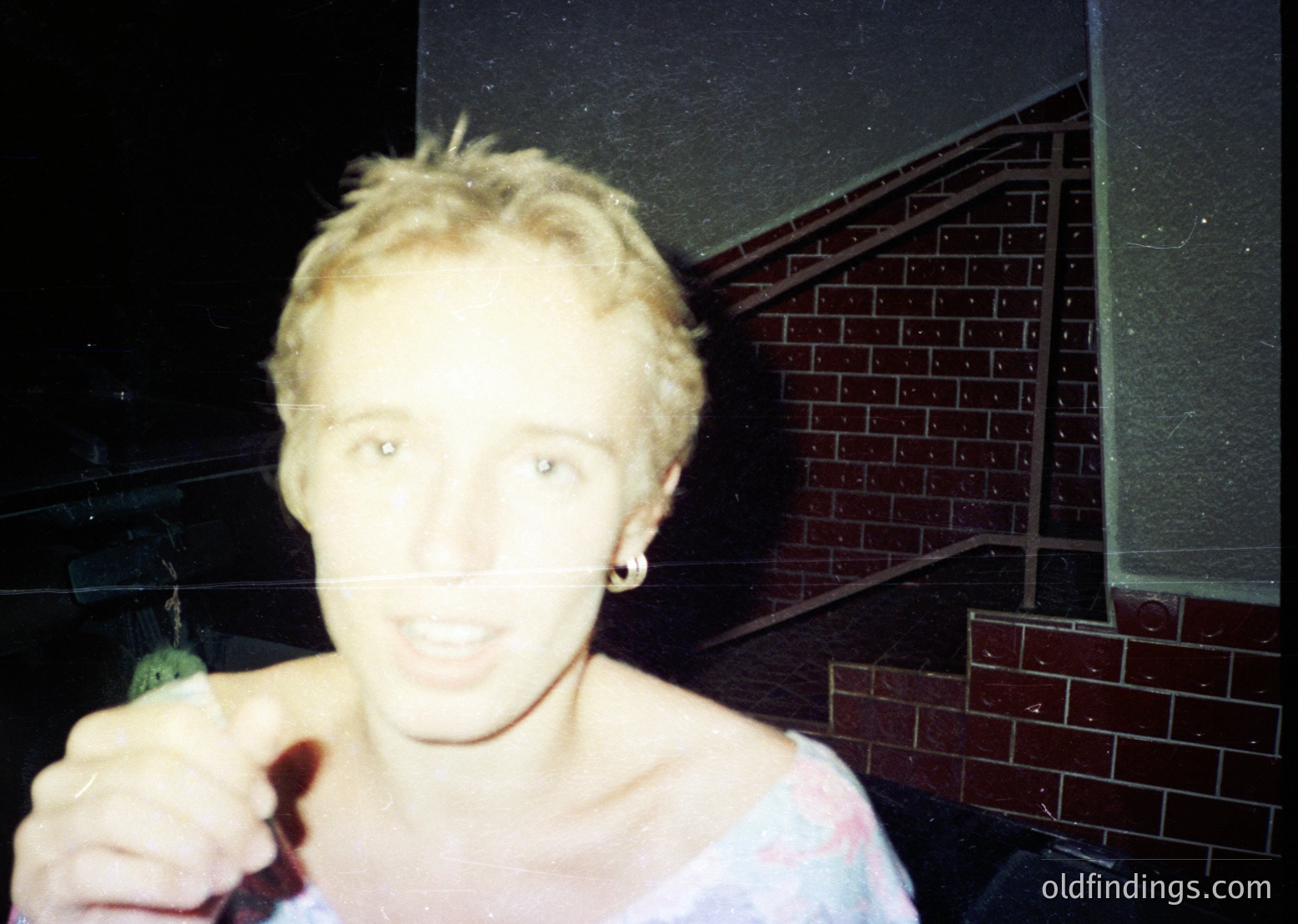 Vintage candid shot of a person holding a green bottle, likely beer, in a dimly lit indoor setting with brick-patterned wall. Short hair, earrings, and casual attire suggest 1990s–2000s era.