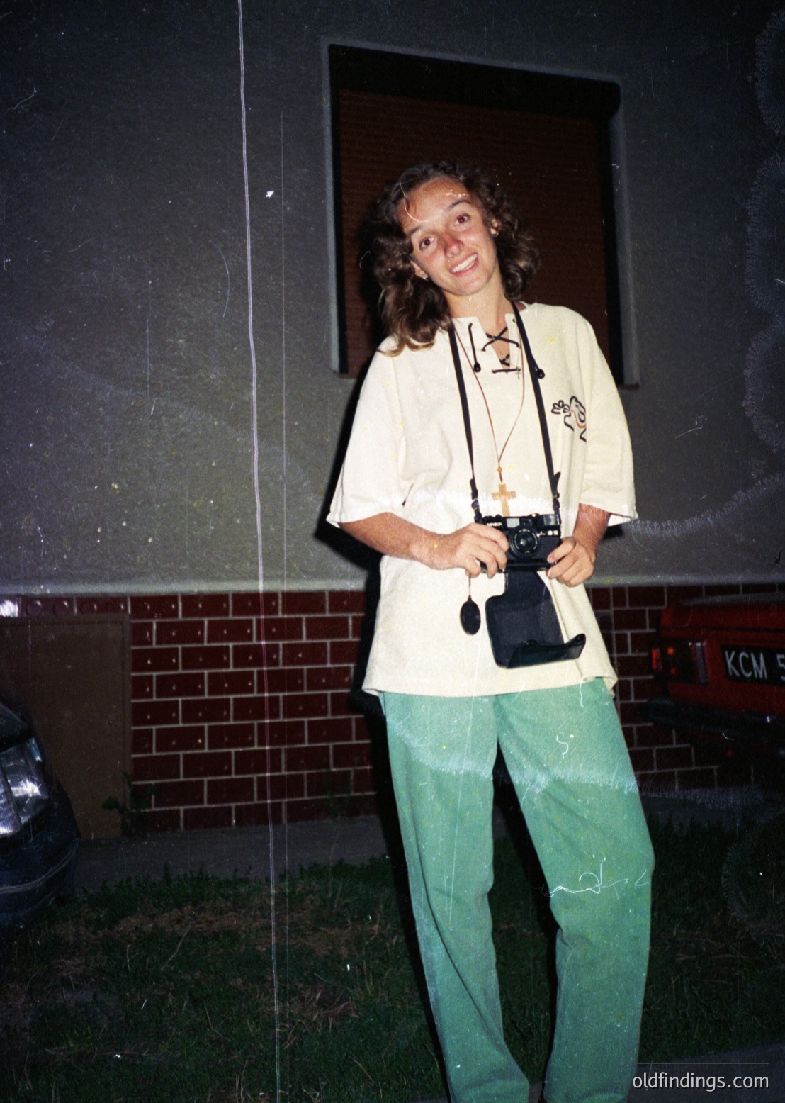 Vintage candid of a person holding a classic film camera, likely a Leica or similar, in front of a brick wall with graffiti. Light-colored sweatshirt with a graphic print, green cargo pants, and a lanyard with a badge or pin. Urban setting, possibly 1970s–1980s.