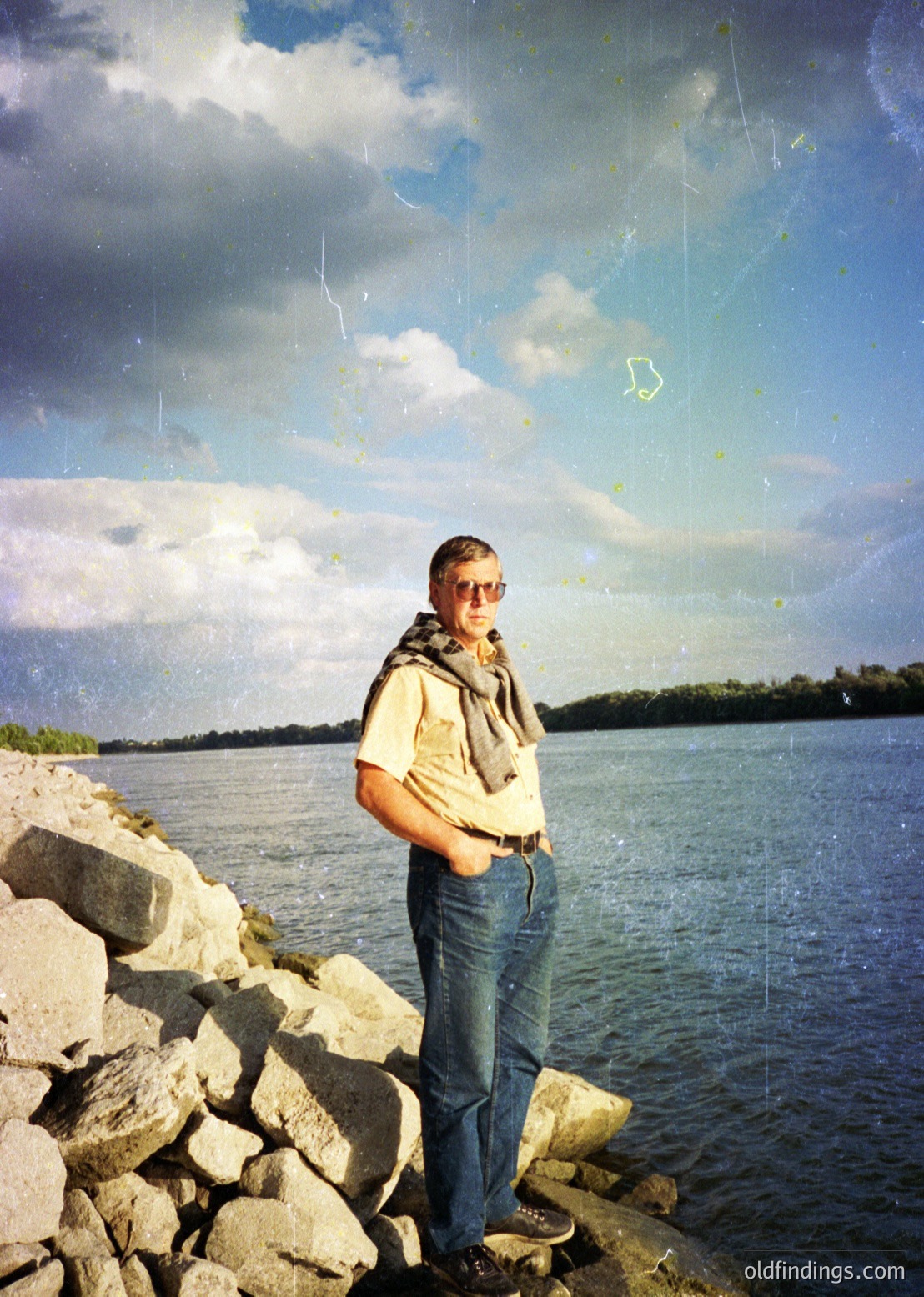 Vintage-style photo of a man in 1970s casual attire—spectacles, vest, jeans—posing by a rocky lakeside. Overcast sky with dramatic clouds and water reflections. Distressed film grain and light scratches enhance nostalgic aesthetic.