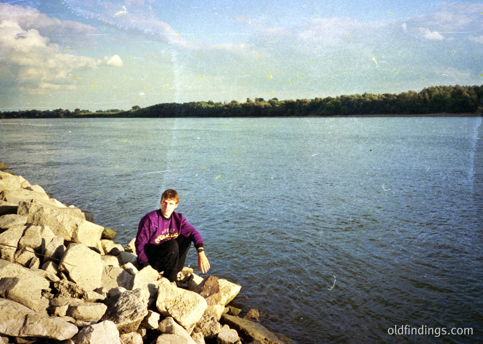 A person sits on rocky terrain beside a calm body of water, holding a small object. The vintage sepia tone suggests mid-20th century (1950s–1970s). Lush forested shoreline in background.