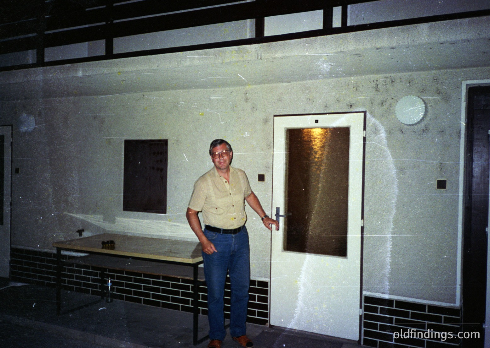 Mid-century institutional hallway with concrete walls and tiled baseboards. Man in casual 1970s attire (short-sleeve button-up, jeans) poses near a frosted glass door. Exposed fluorescent lighting and a blank wall plaque suggest a utilitarian or educational setting.
