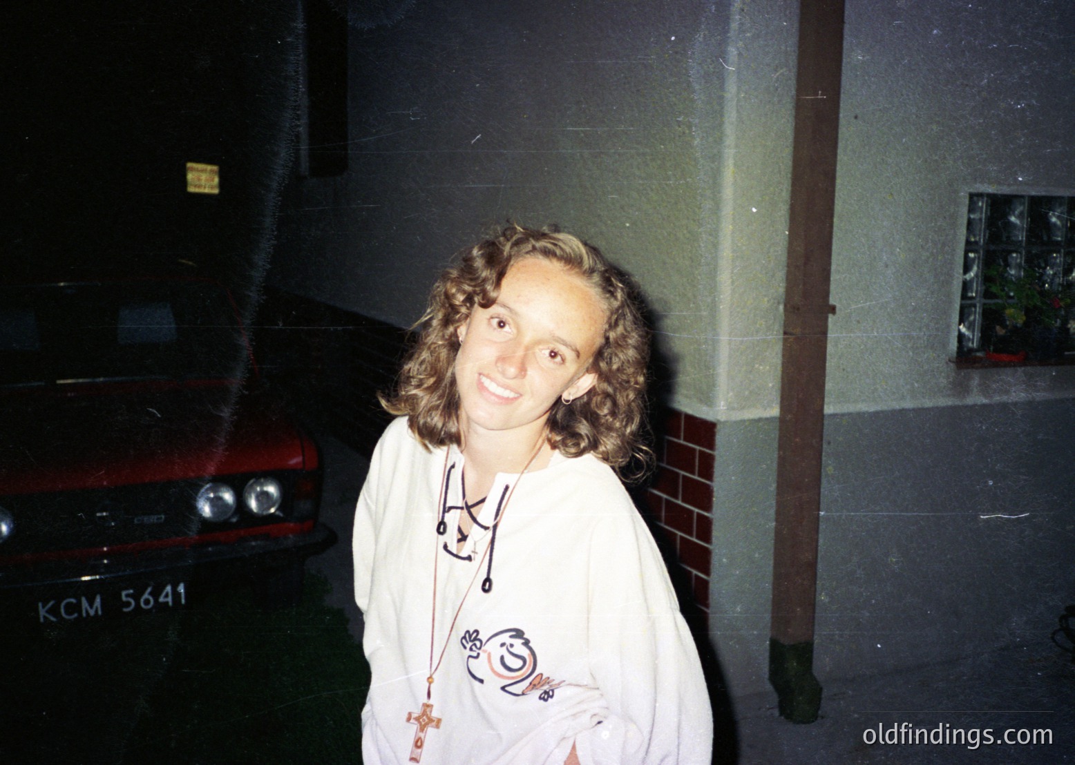 Vintage candid of a person in a 1980s-style hooded sweatshirt with cartoon graphics, standing beside a red classic car (KCM 5641) in dim indoor lighting. Brick wall and potted plant in background suggest a garage or basement setting.