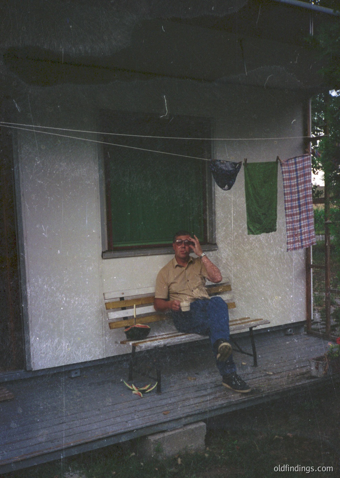 Man in casual 1970s attire rests on a wooden bench outside a weathered concrete building. Green window, green towel, and checkered cloth hung to dry. Overcast lighting suggests midday.