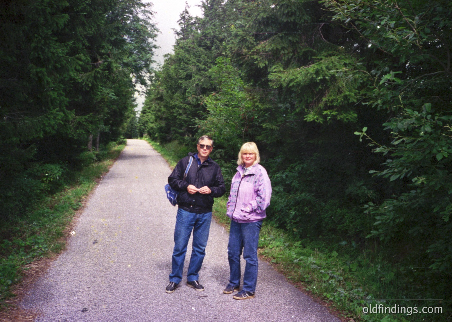 Two individuals pose on a paved forest path, surrounded by dense greenery. The man wears a dark jacket, jeans, and a backpack; the woman wears a pink jacket, jeans, and sneakers. Likely taken in the late 2000s or early 2010s, this image captures a serene outdoor moment.