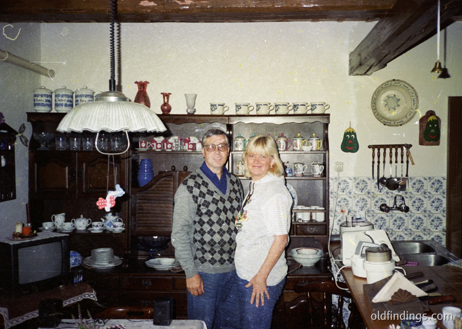 Vintage kitchen interior featuring wooden cabinets, blue-and-white patterned tiles, and ceramic dishes. Two individuals pose in 1970s-style attire—man in checkered sweater, woman in white blouse. Decorative items include vintage lamps, vases, and wall art. Warm, lived-in ambiance.