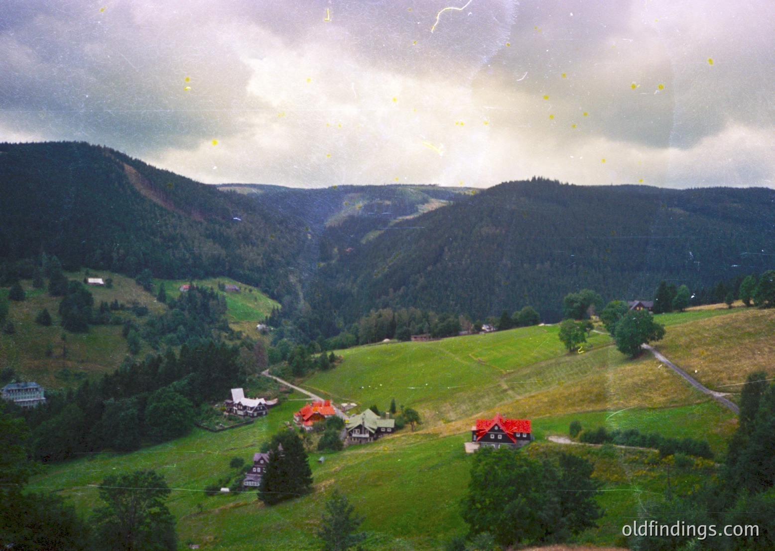 Aerial view of alpine valley with dense forest-covered slopes and scattered farmhouses. Green pastures dominate the foreground, with a winding road cutting through. Clouds obscure the upper sky, hinting at impending rain. Likely European alpine region, mid-20th century.