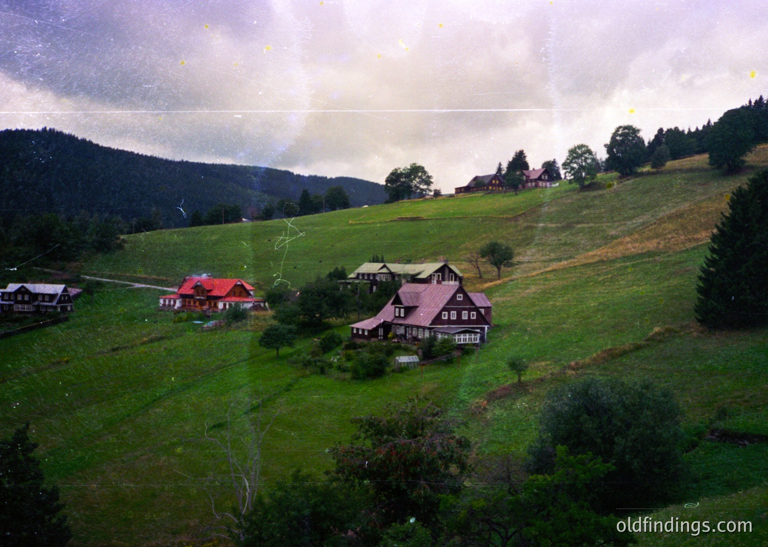 Aerial view of a rural Alpine village with clustered wooden chalets on rolling green hills. Central building features a red-tiled roof and white trim, surrounded by dense forest and misty peaks. Likely European alpine region, possibly Austria or Switzerland.