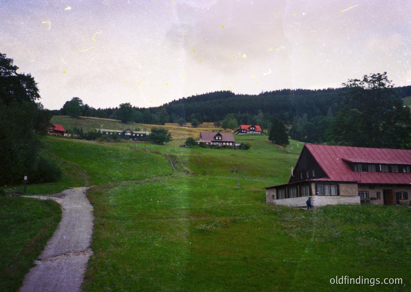 Vintage rural landscape featuring a winding gravel road leading through lush green meadows. Mid-20th century farmhouses with red roofs and white walls dot the rolling hills, surrounded by dense forest. Overcast sky enhances serene, pastoral atmosphere. Likely Eastern European countryside, 1950s-1970s.