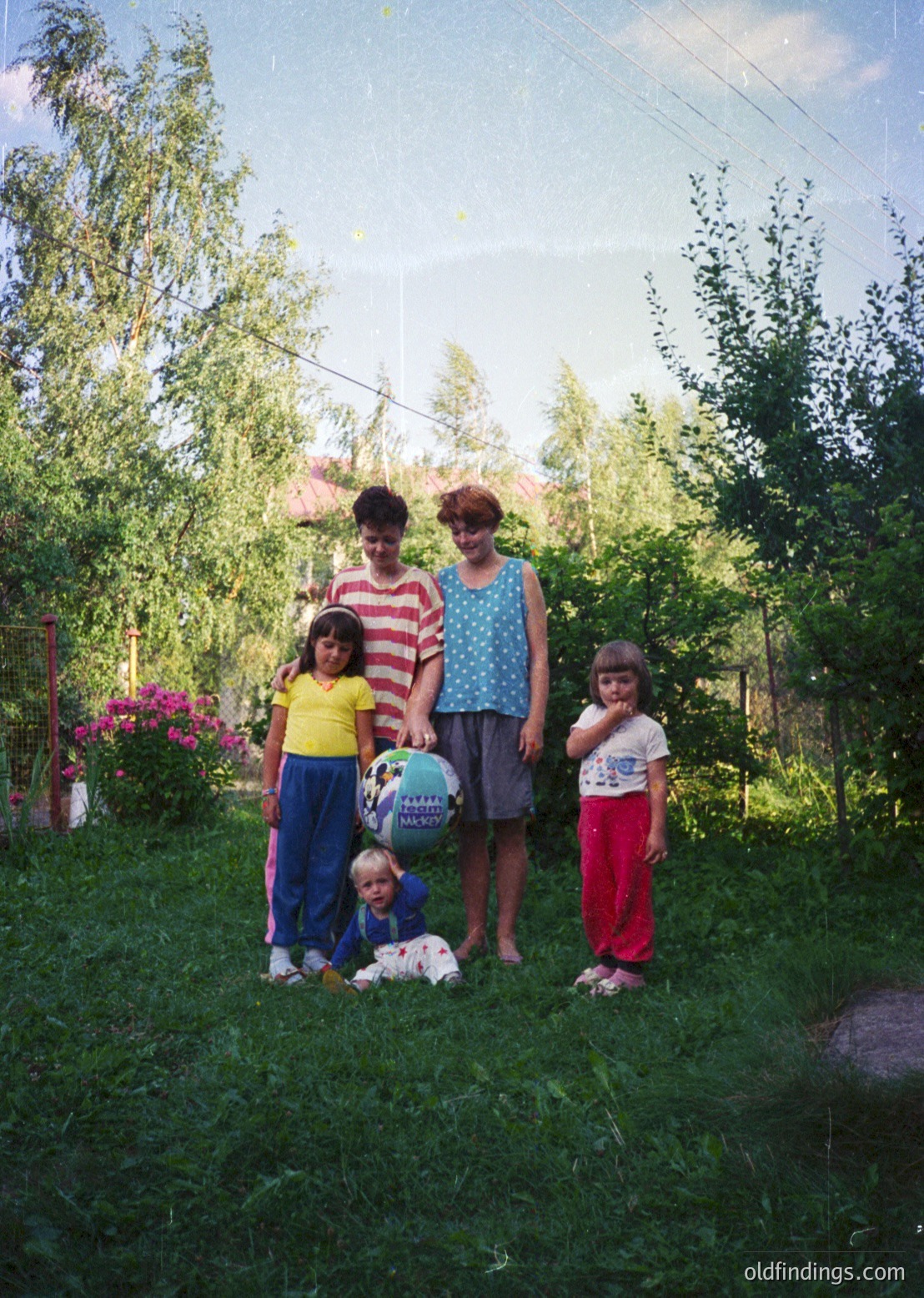 Family portrait in a lush garden setting, likely Eastern Europe, 1970s–1980s. Four individuals pose on grass with vibrant greenery, blooming flowers, and mature trees. Adults wear patterned blouses and striped shirts; children in bright, practical clothing. A child holds a round, textured ball with Cyrillic script. Soft lighting suggests daytime, outdoor leisure.