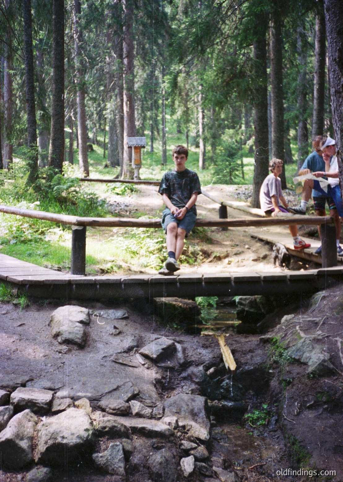 Wooden boardwalk bridge in a dense forest, with three young men posing. One sits on the railing, another stands on the bridge, and the third leans on a rock beside the waterfall. Mid-20th century outdoor recreation attire ( )