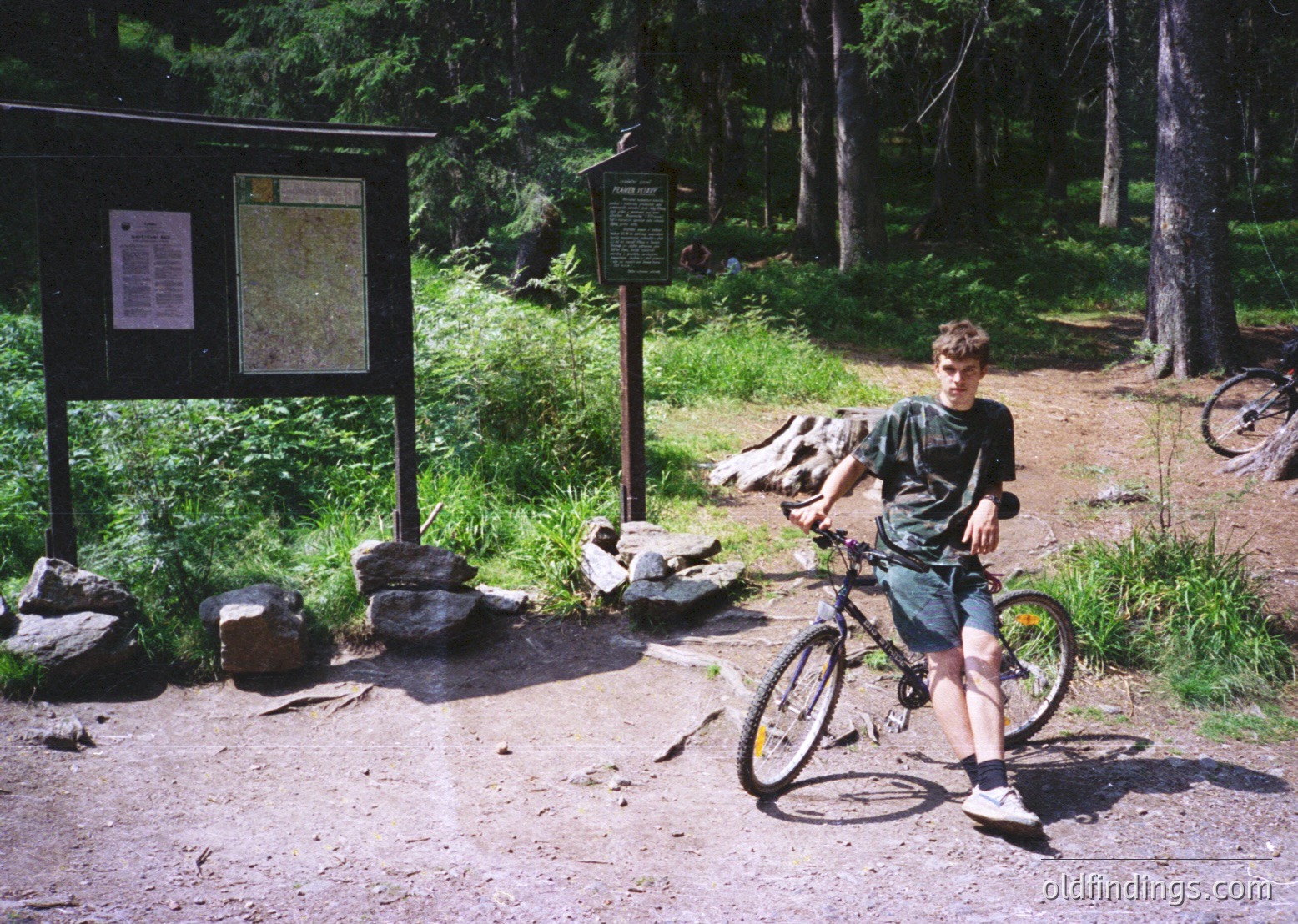 A young person poses with a classic mountain bike at a forested trailhead, surrounded by informational signs and a map. The scene suggests a mid-1990s to early 2000s outdoor adventure, likely in a temperate forest region.