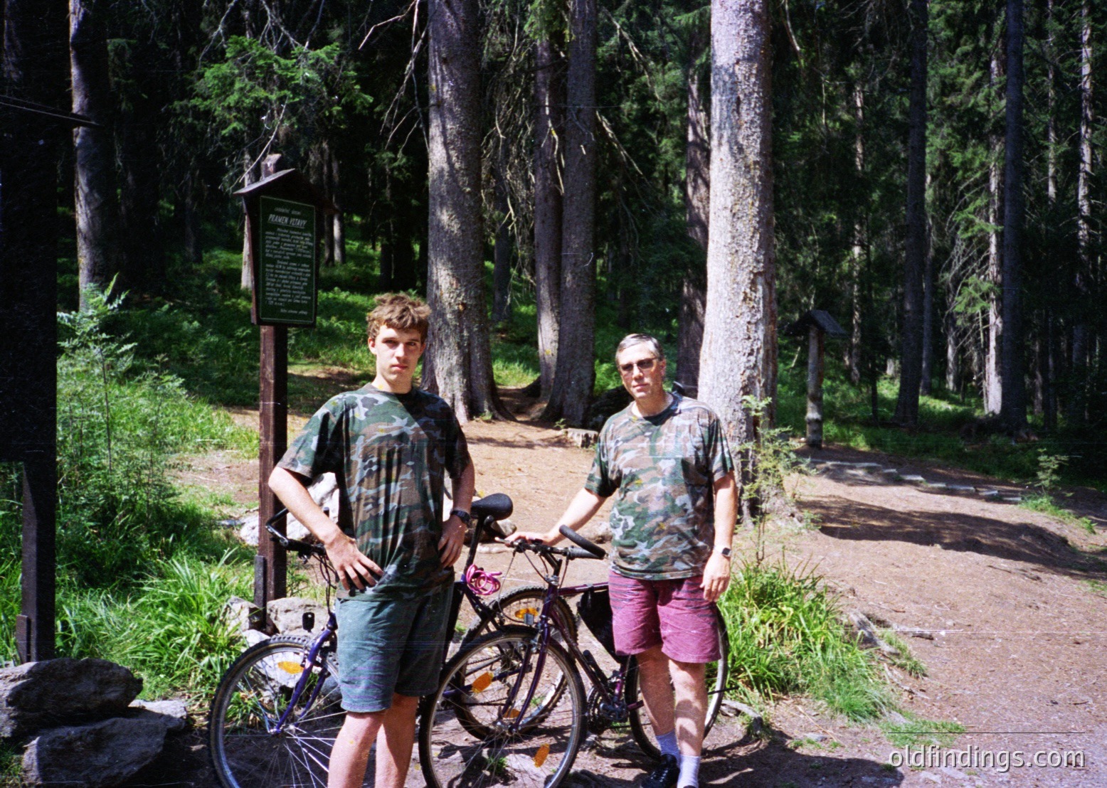 Two men pose with bicycles in a forested area, likely a park or nature trail. The man on the left wears a tie-dye shirt and shorts, while the other sports a floral-patterned shirt and pink shorts. Bikes lean against a wooden signpost. Dense pine trees frame the scene, suggesting a temperate forest setting. Style hints at 1970s outdoor recreation.