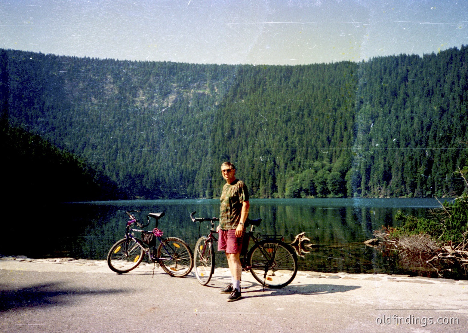 Retro-style photo of a man posing with two bicycles beside a serene alpine lake, framed by dense coniferous forests and snow-capped peaks. Likely 1980s–1990s European mountain region.