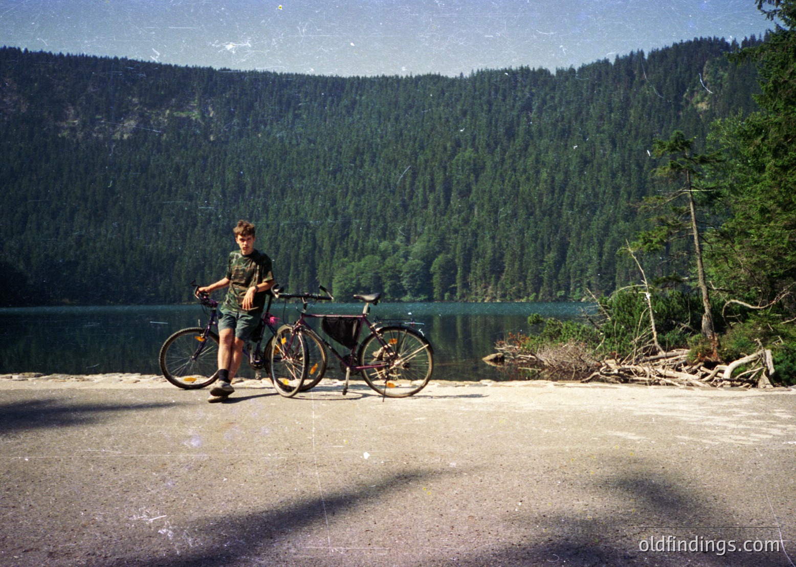 Vintage-style photo of a person posing with a classic mountain bike beside a serene alpine lake, surrounded by dense coniferous forests. The individual wears a green top and shorts, typical of mid-20th-century outdoor fashion. The scene suggests a recreational cycling trip in a mountainous region, likely the European Alps or similar terrain.