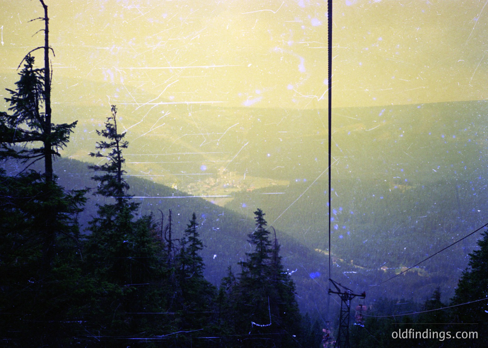 Contrast-rich alpine forest scene viewed through a fogged or scratched glass surface, distorting clarity. Dense evergreen trees frame a valley below, with misty mountain ridges fading into a hazy horizon. Likely captured from an elevated vantage point, possibly a ski lift or observation deck.