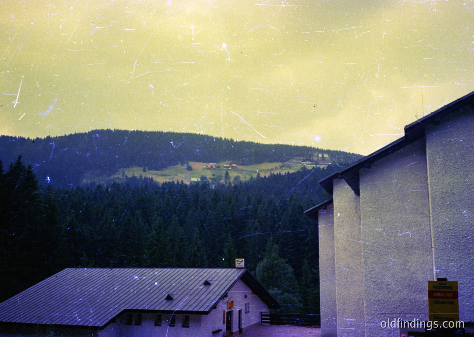 Vintage alpine resort scene with aged photo filter. Dense evergreen forest slopes into rolling hills under hazy golden light. Mid-century concrete buildings with flat roofs and minimalist design suggest a 1960s-70s European ski lodge or mountain retreat.