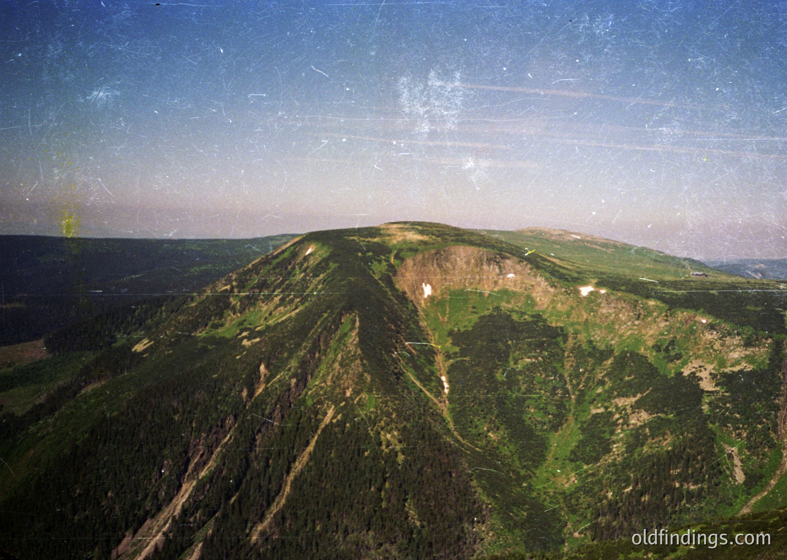 Aerial view of rugged alpine terrain with steep slopes and dense forestation. The image captures a mix of rocky outcrops and lush greenery, likely part of a high-altitude region. The sky shows faint contrails, suggesting recent aircraft passage. The composition highlights natural beauty and potential for adventure tourism.