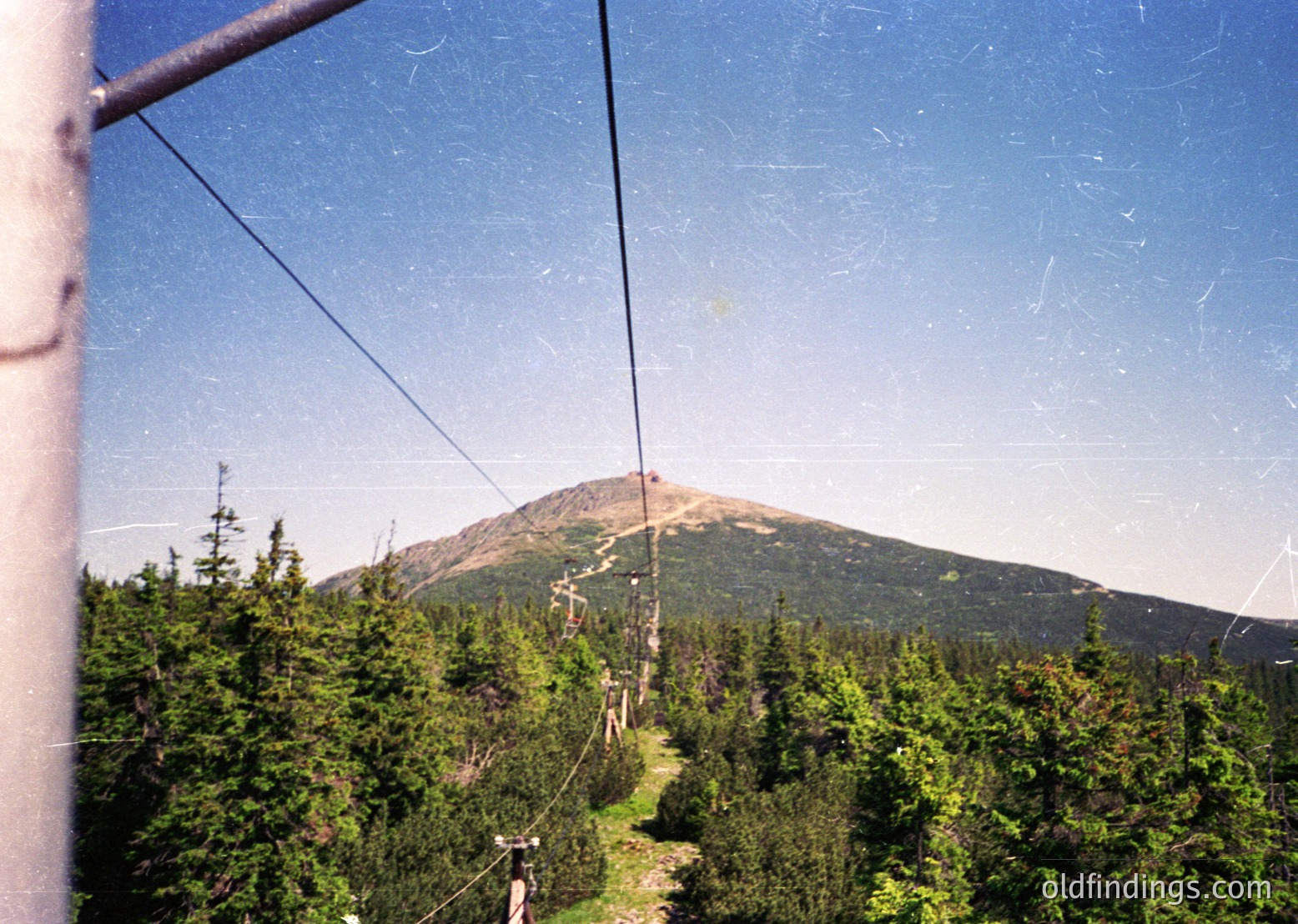 Vintage aerial tramway cable view of forested slopes leading to a snow-capped peak, framed by metal support structure. Dense coniferous forest dominates the midground under clear skies. Likely Eastern European alpine region, 1970s-1990s era.