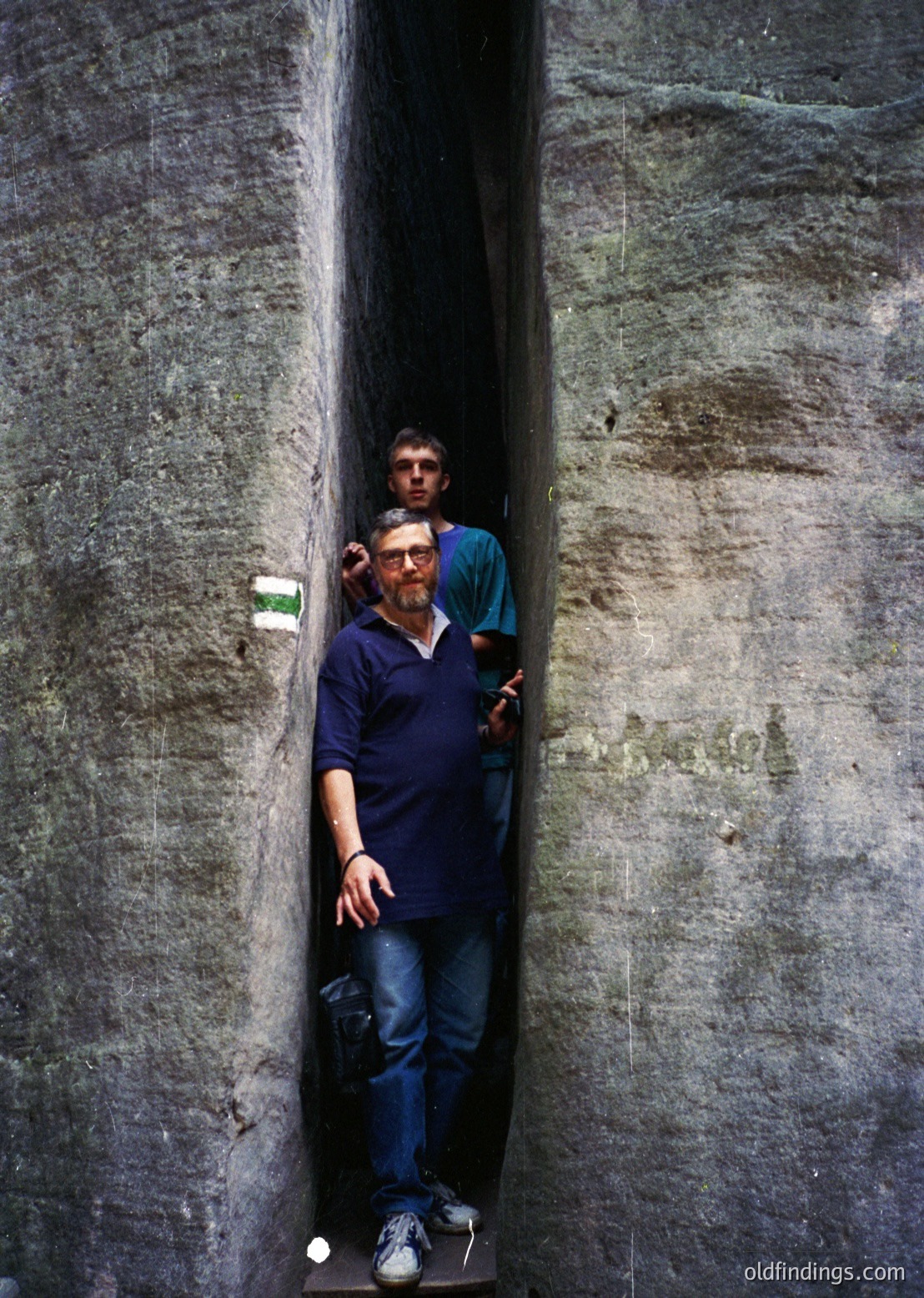 Two men pose in a narrow, weathered concrete tunnel. The man in front wears a blue polo shirt, jeans, and sneakers; the other, in teal, stands behind him. The tunnel’s rough texture and lighting suggest an industrial or urban setting. Likely 2000s–2020s, modern era.