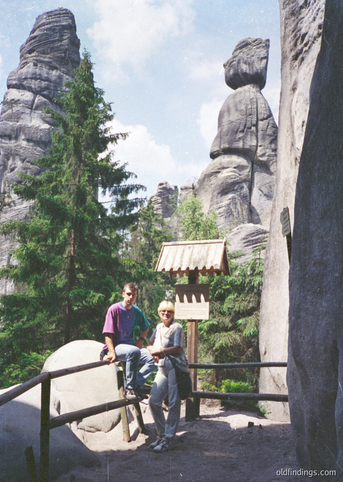 Two individuals pose on a wooden bridge near the famous **Bastei** rock formations in **Sächsische Schweiz National Park**, Germany. The scene captures iconic sandstone cliffs, lush greenery, and a traditional wooden shelter ( ).
