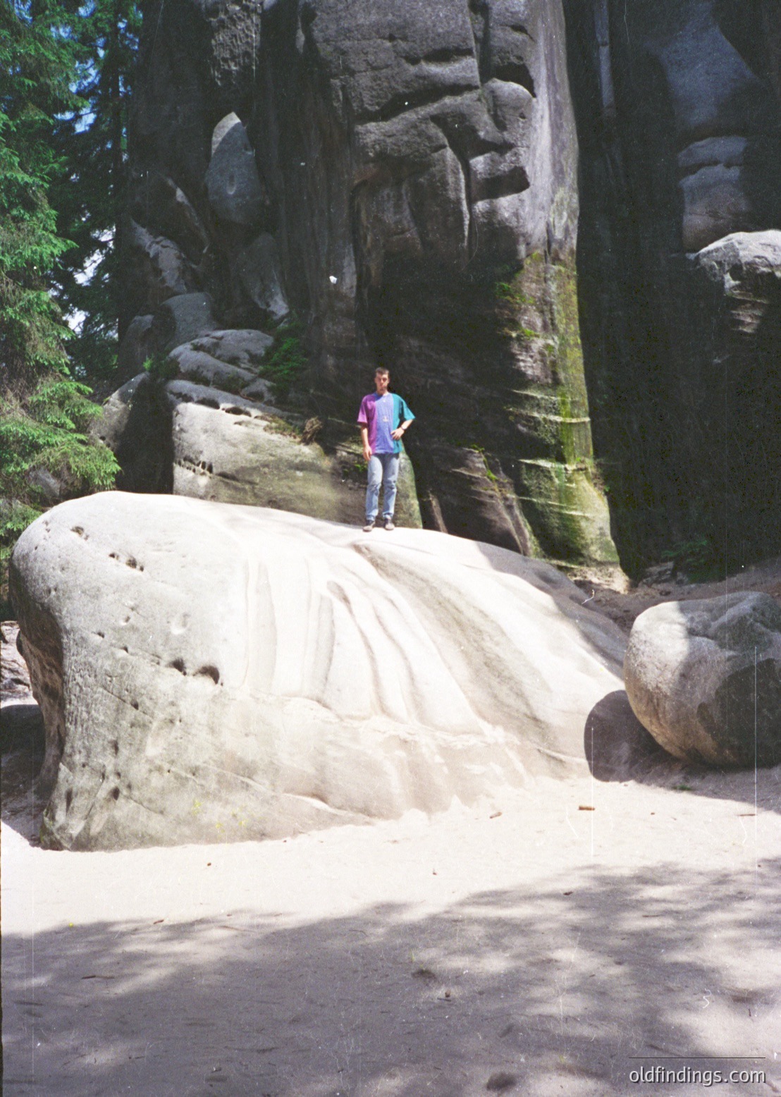 A person stands on a smooth, rounded boulder in a rocky canyon, surrounded by towering sandstone formations. The scene captures striking vertical rock walls with moss-covered textures, suggesting a natural limestone landscape. The lighting highlights dramatic contrasts between light and shadow, indicative of a sunny day.