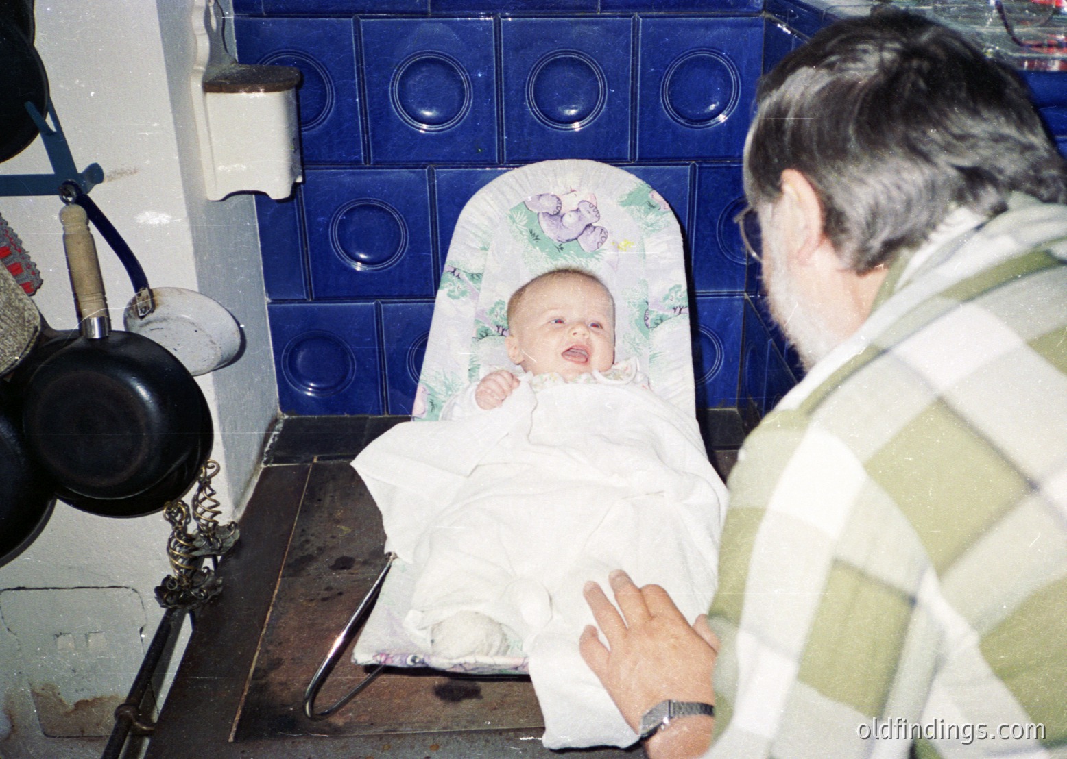 Vintage kitchen scene featuring a baby in a highchair with floral-patterned padding, swaddled in white. Adult male in checkered shirt interacts with infant. Blue ceramic tiles with circular motifs line the wall. Cast-iron pot and vintage stove visible. Likely Eastern European domestic setting, 1980s–1990s.