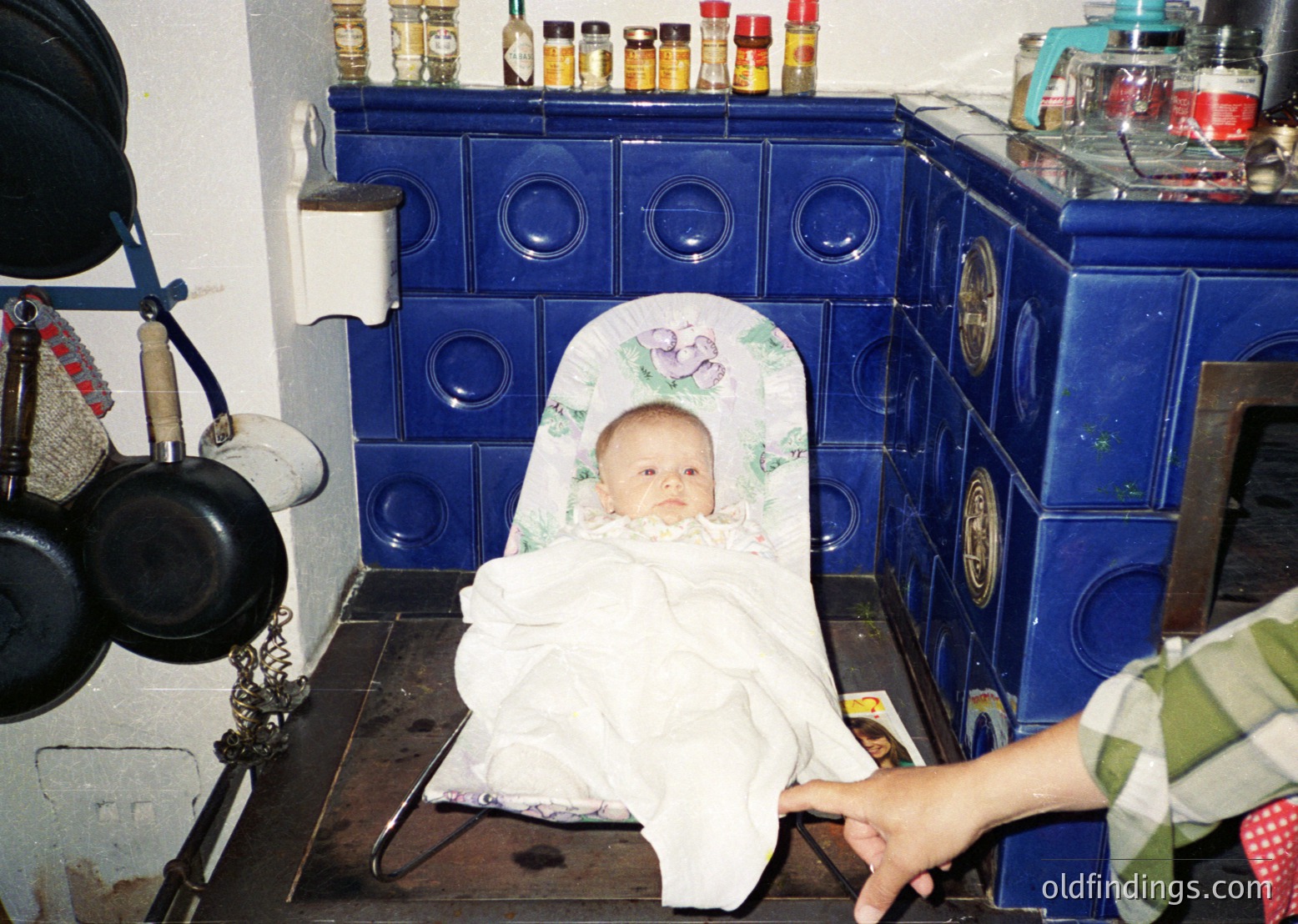Vintage kitchen scene featuring a baby seated on a blue-tiled stove, wrapped in a floral-patterned cloth. Surrounding items include vintage cookware (pots, kettles) and spice jars. Warm, nostalgic domestic atmosphere. Likely Eastern European, mid-20th century.