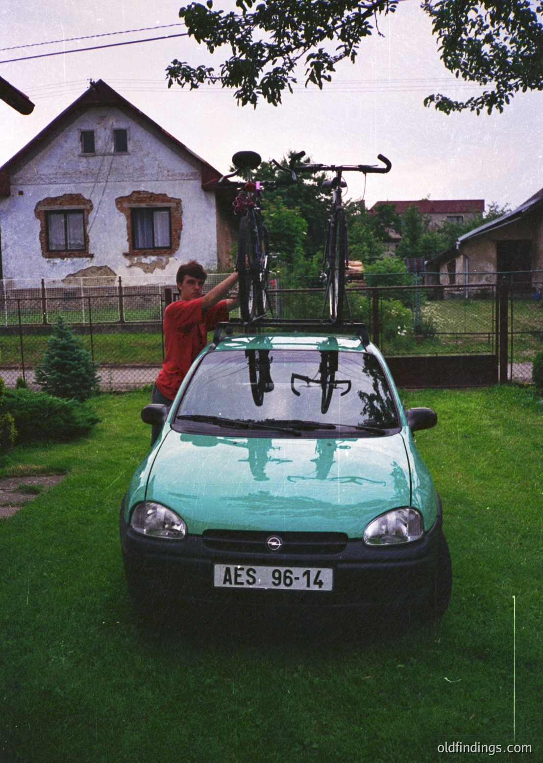 Green hatchback (likely a Renault Twingo) with roof-mounted bike rack carrying two bicycles, parked on grassy residential lawn. Man in red shirt secures bike. Whitewashed house with aged plaster and red-tiled roof in background. Suburban setting, likely Eastern Europe, 1990s–2000s.