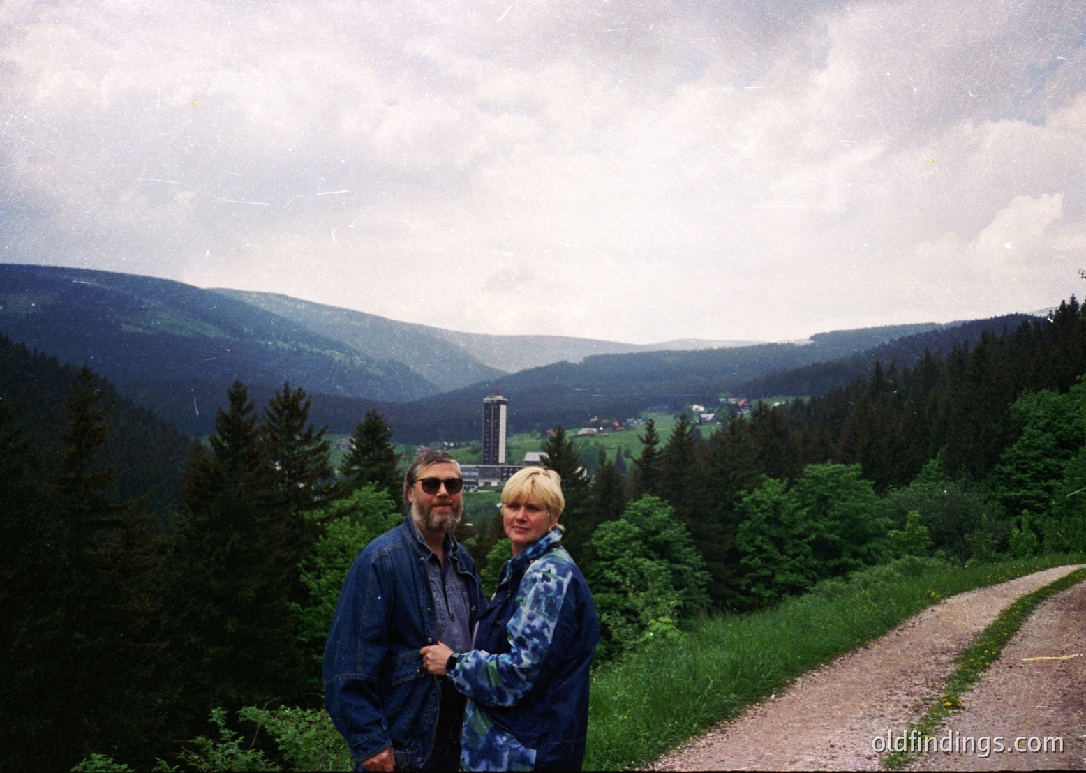 Two individuals pose on a winding forest path surrounded by dense evergreen trees, set against rolling hills and a distant tower. Clothing suggests 1980s–1990s outdoor fashion ( ).