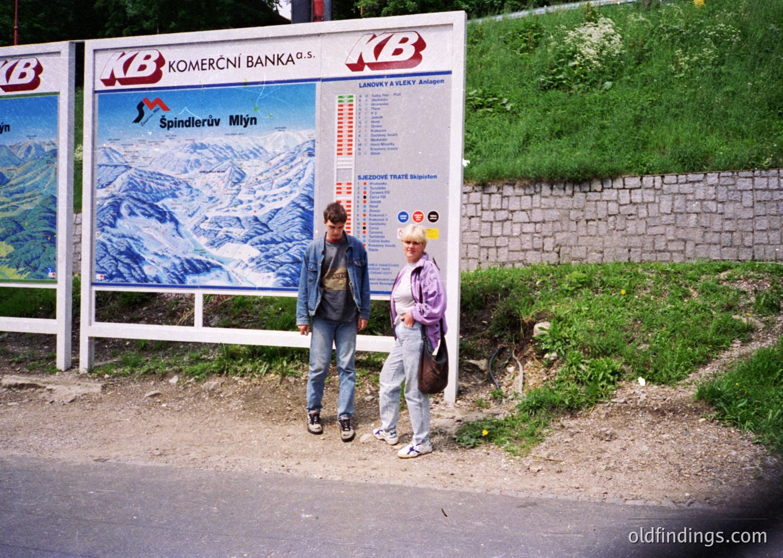 Two individuals pose beside a vintage Czech bank billboard advertising ski resort "Špindlerův Mlýn," featuring alpine imagery. The sign includes lift schedules and pricing. Likely taken in the 1990s in the Czech Republic.