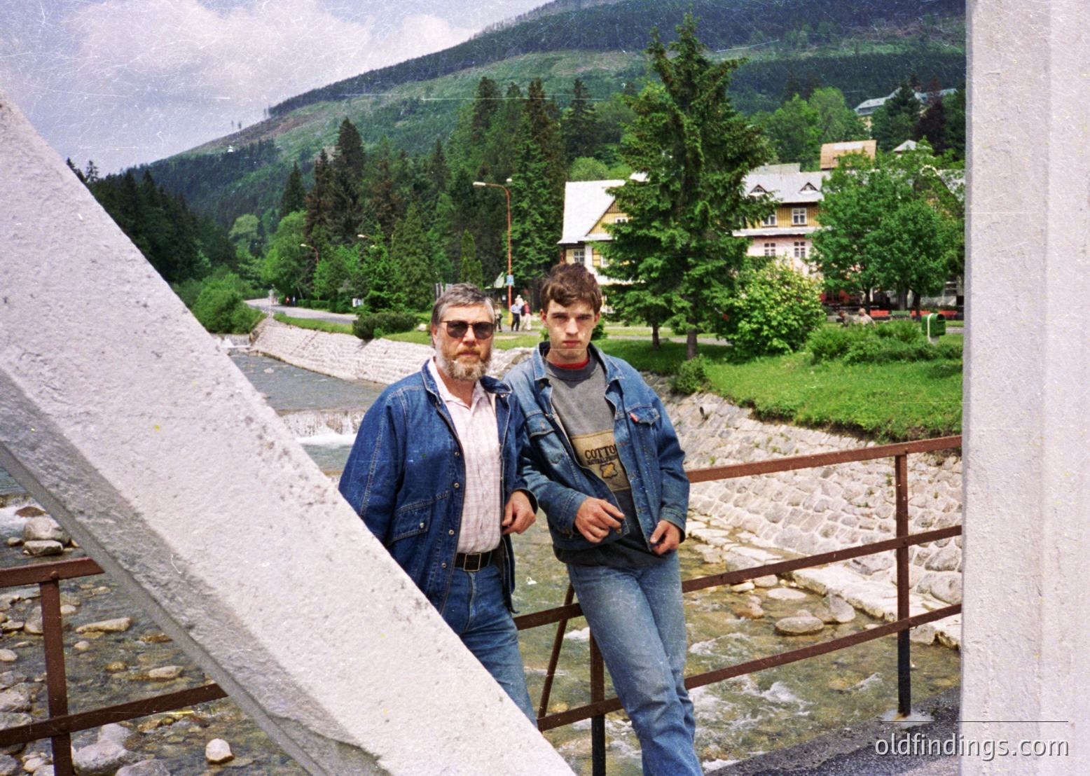 Two men pose on a stone bridge overlooking a lush alpine valley, framed by a concrete structure. The older man wears a denim jacket and sunglasses; the younger sports a graphic tee and jeans. Wooden chalet-style buildings and dense pine forests surround a winding road. Likely taken in the 1990s–2000s in a European alpine region.